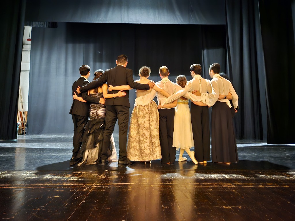 Otto artisti in abiti da scena, abbracciati sulla ribalta di un teatro, visti da dietro.