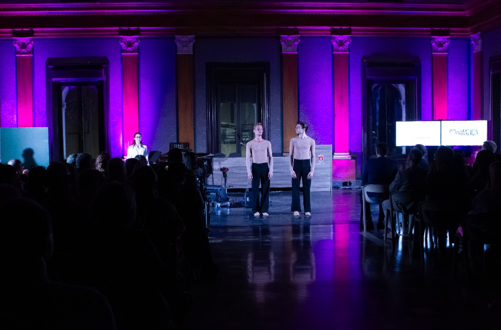 Due performer in sala storica illuminata dal magenta; sullo schermo il logo CROMATICA, pubblico in sala.