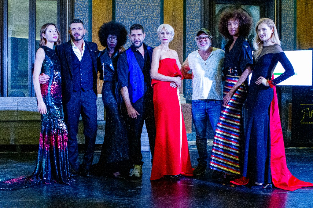 Gruppo di otto persone in posa sul palco — ospiti e performer in abiti da sera e casual, ambiente luminoso.