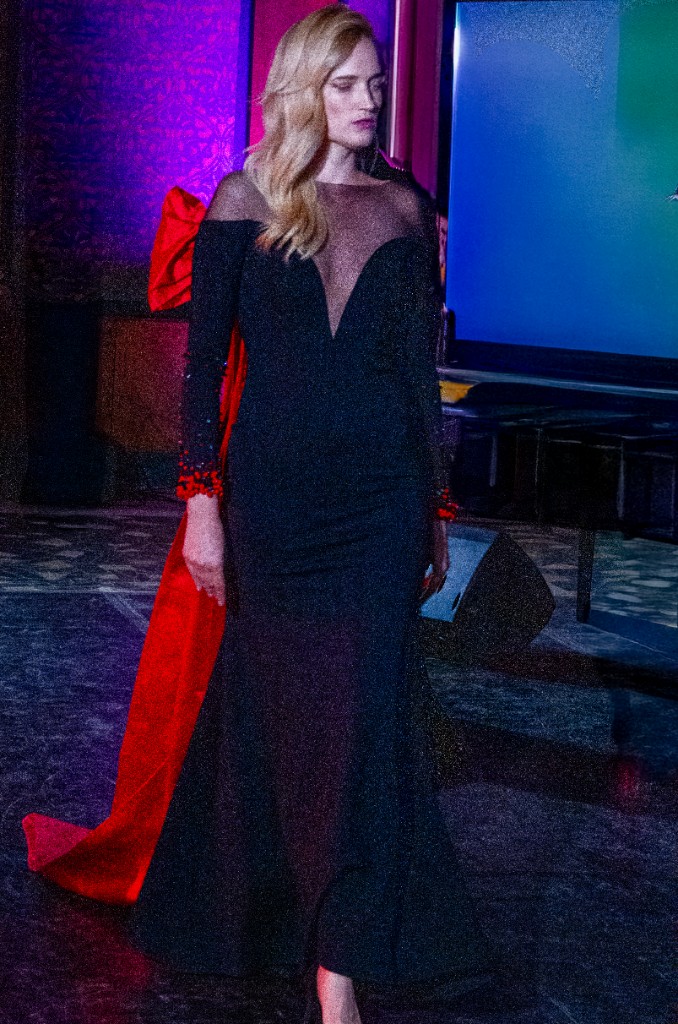 Modella in lungo abito nero con maniche decorate e grande fiocco rosso sulla schiena, in passerella.