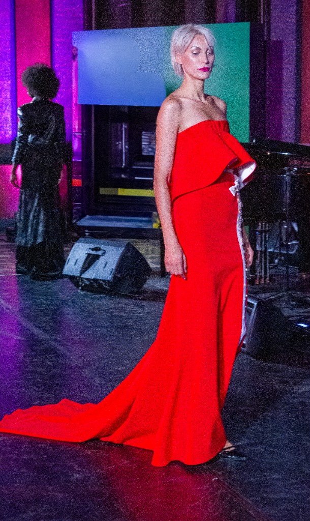 Modella bionda in lungo abito rosso con ruche e strascico, passerella con luci viola e verdi.