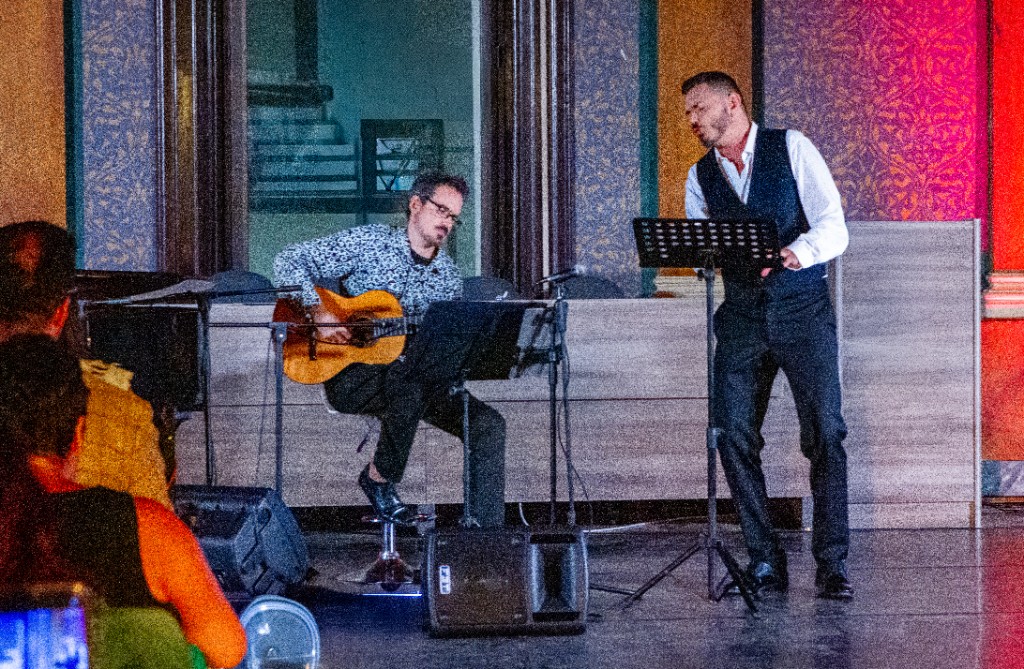 Musica dal vivo: chitarra acustica e voce solista in sala con carta da parati damascata; in primo piano il pubblico.