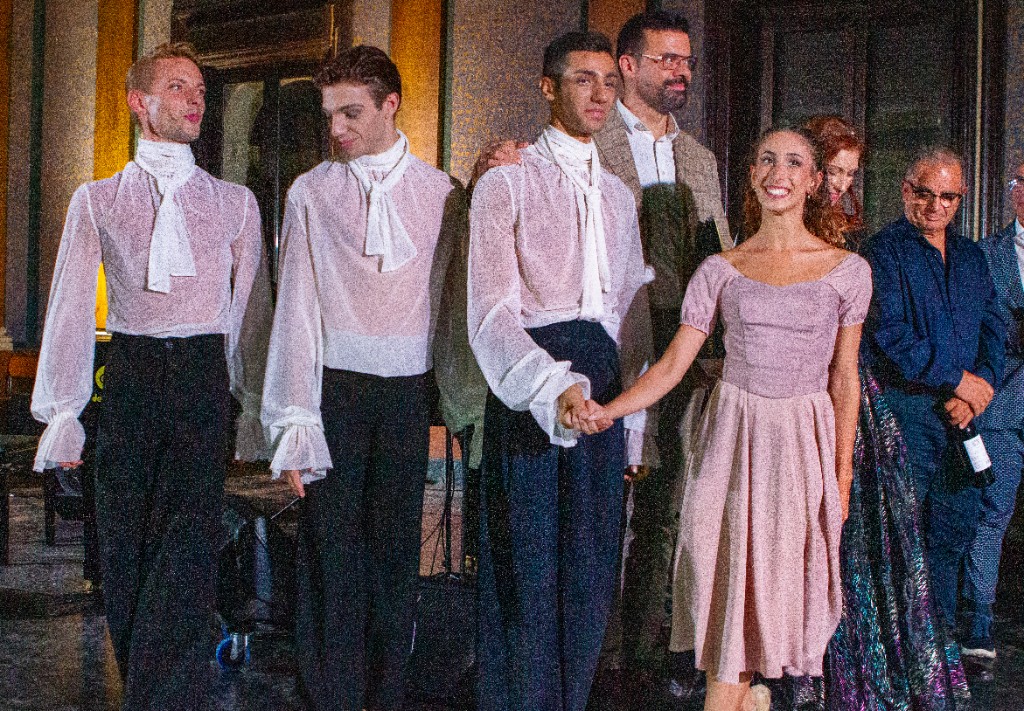 Compagnia di danza in costume e organizzatori sul palco dopo lo spettacolo, atmosfera conviviale.