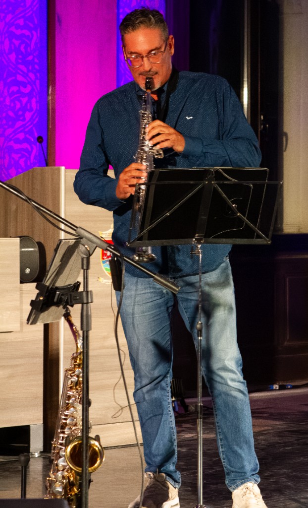 Musicista con sassofono soprano su palco in legno, leggio e luci viola sul fondale.