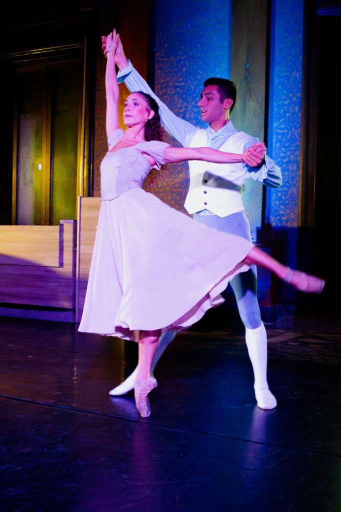 Coppia di danzatori classici in equilibrio sul palco, luci blu e fondale decorato.