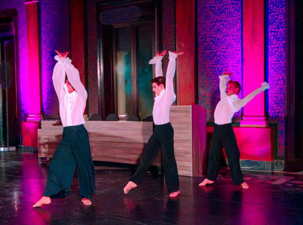 Tre danzatori in camicia bianca e pantaloni neri in movimento espressivo, palco con luci magenta.