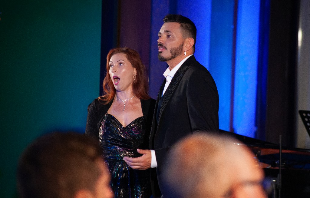 Soprano e tenore in abito da sera durante un duetto lirico dal vivo, pubblico in primo piano.
