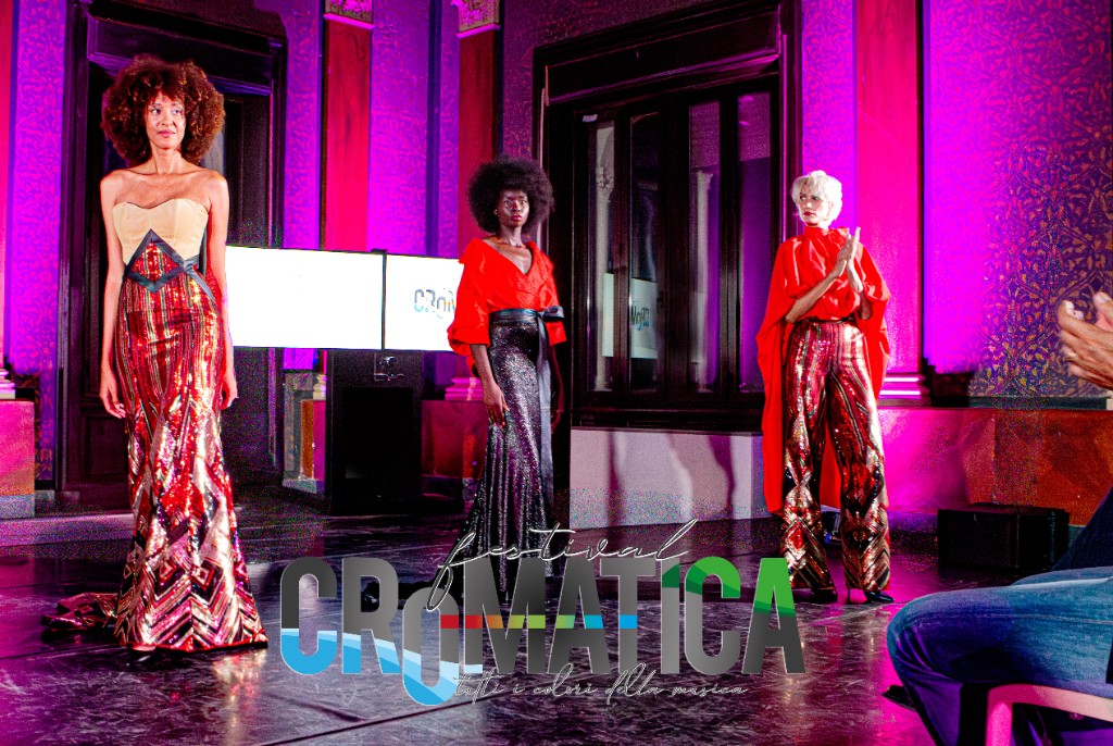 Tre modelle in passerella con look da sera e palette rosso-oro, ambientazione da sfilata CROMATICA Festival.