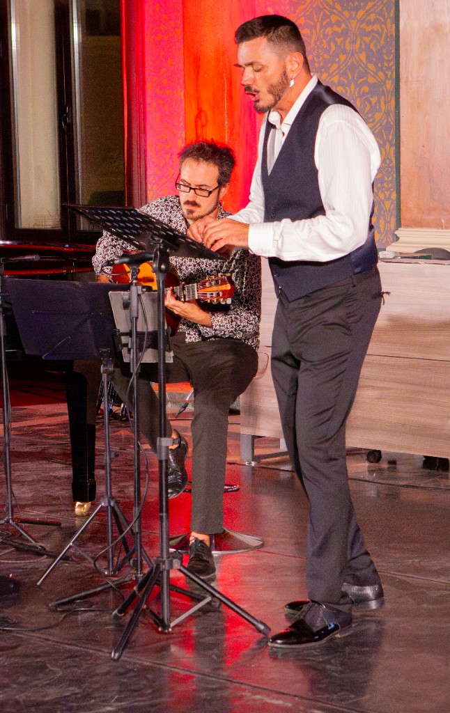 Performance musicale dal vivo: voce con gilet scuro e musicista alla chitarra acustica, luci calde sul palco.